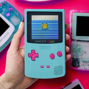 May include: A teal Game Boy Color handheld console with pink buttons and a screen displaying a Pikachu graphic. The text "GAME BOY COLOR" and "Nintendo" are visible. Other Game Boy consoles are in the background.