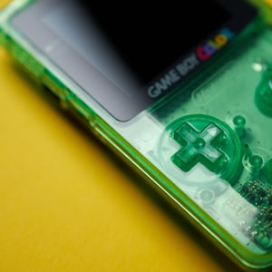 Op de afbeelding: Een draagbare Game Boy Color spelcomputer met een doorschijnende neon-groene plastic behuizing. Het apparaat bevat een richtingspad, twee ronde actieknoppen en een zwarte schermrand met het Game Boy Color logo in het wit gedrukt.
