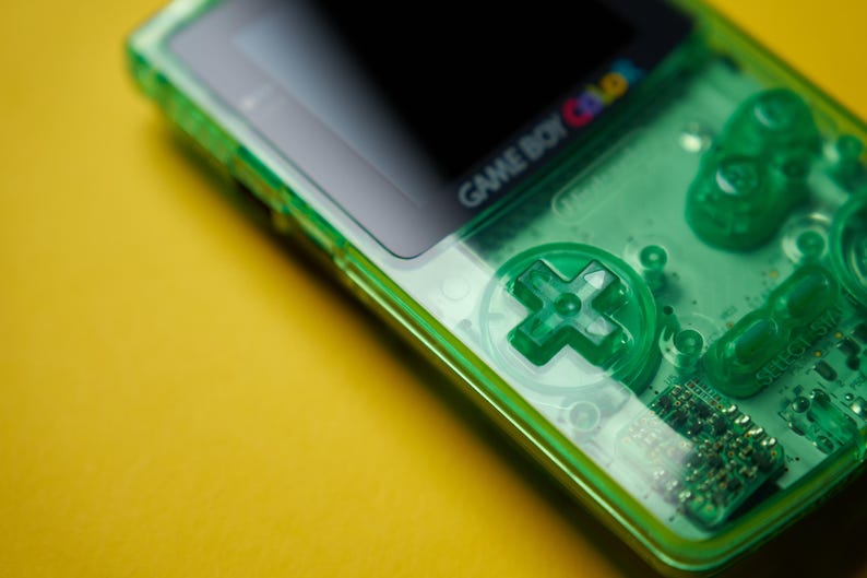 Op de afbeelding: Een draagbare Game Boy Color spelcomputer met een doorschijnende neon-groene plastic behuizing. Het apparaat bevat een richtingspad, twee ronde actieknoppen en een zwarte schermrand met het Game Boy Color logo in het wit gedrukt.