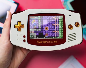 Peut inclure: Une console de jeu portable Game Boy Advance blanche et rouge avec des finitions dorées. L'écran affiche un jeu vidéo avec un personnage dans un monde violet et vert. La console a un bouton rouge et blanc sur le côté gauche et un bouton doré sur le côté droit. Le texte "GAME BOY ADVANCE" est imprimé en bas de la console.
