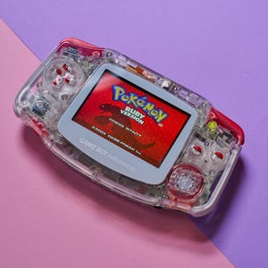 May include: A clear, red, and white Game Boy Advance SP handheld gaming console with a translucent shell. The screen displays the title screen for the game "Pokémon Ruby Version" with the text "Press Start" and "© 2003 GAME FREAK Inc."