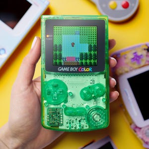Op de afbeelding: Een limoengroene doorschijnende Game Boy Color handheld spelcomputer met een zichtbare interne printplaat, een richtingspad, twee actieknoppen en een scherm dat een gepixeld personage in een groene spelomgeving weergeeft.