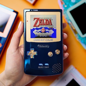 Puede incluir: Consola portátil Game Boy Color azul marino que muestra "The Legend of Zelda: Link's Awakening DX". La consola tiene detalles dorados en los botones y una cruceta direccional. Otras consolas son visibles en el fondo.