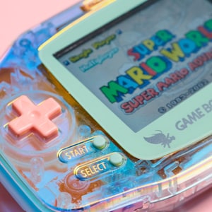 May include: A translucent blue and green handheld games console with a pink cross button, start and select buttons, and a screen displaying the text "Super Mario World Super Mario Advance &copy; 1985-2001 Game Boy Advance".