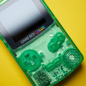 Op de afbeelding: Een doorschijnende limoengroene Game Boy Color handheld spelcomputer met een zichtbare interne printplaat, een richtingspad, A- en B-knoppen, en Start- en Select-knoppen. Het schermframe toont het Game Boy Color logo.
