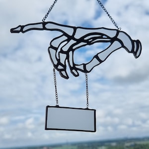 May include: A stained glass skeleton hand with a blank sign hanging from a chain against a blue sky with white clouds.