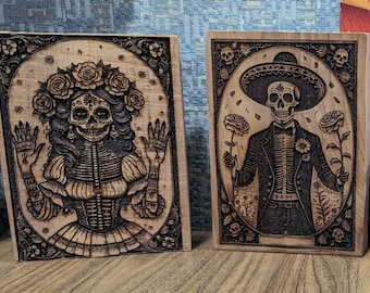 May include: Two wooden plaques with laser-etched images of skeletons in traditional Mexican Day of the Dead attire. The woman is wearing a floral crown and a dress, while the man is wearing a suit and a sombrero.
