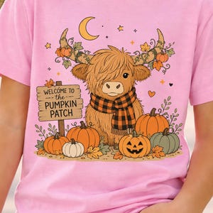 May include: A light pink short-sleeve t-shirt featuring a graphic of a fluffy highland cow wearing a plaid scarf. The design includes pumpkins, autumn leaves, a crescent moon, and a wooden sign that reads Welcome to the Pumpkin Patch.