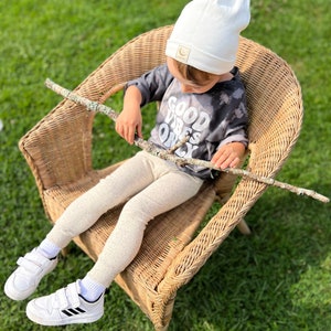 May include: A child wearing a white beanie with a moon logo sits in a wicker chair. The child is wearing a grey shirt with the text "Good Vibes Only" and beige leggings. The child is holding a stick in their hands.