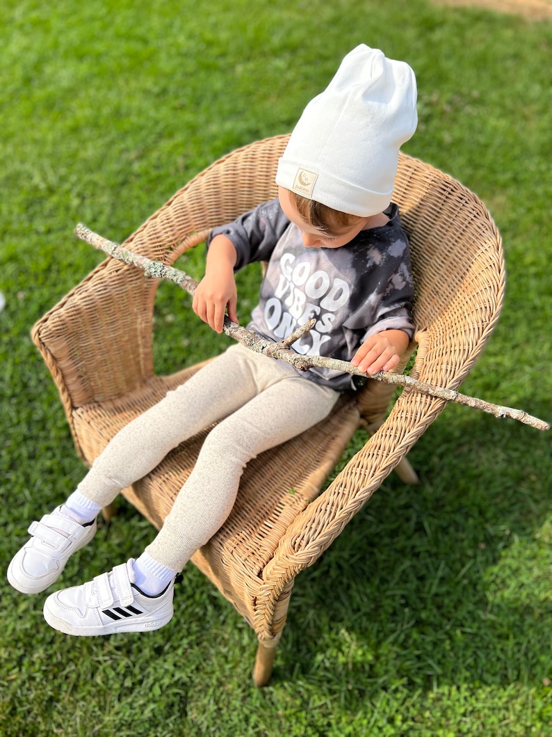 May include: A child wearing a white beanie with a moon logo sits in a wicker chair. The child is wearing a grey shirt with the text "Good Vibes Only" and beige leggings. The child is holding a stick in their hands.