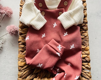 May include: A red and white baby onesie with a star pattern. The onesie is in a wicker basket with a white long-sleeved shirt on top.