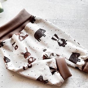 May include: Baby pants with a white background and a pattern of woodland animals, including foxes, rabbits, badgers, and boars. The cuffs and waistband are a solid brown color. The pants are made of a soft fabric.