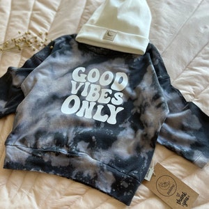 May include: Black and white tie-dye sweatshirt with the words "GOOD VIBES ONLY" in white. The sweatshirt is paired with a white beanie.