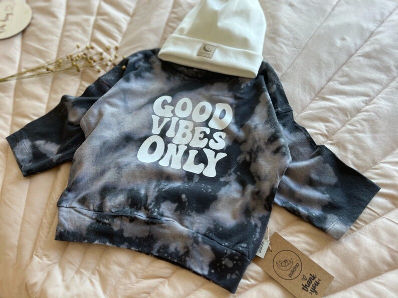 May include: Black and white tie-dye sweatshirt with the words "GOOD VIBES ONLY" in white. The sweatshirt is paired with a white beanie.