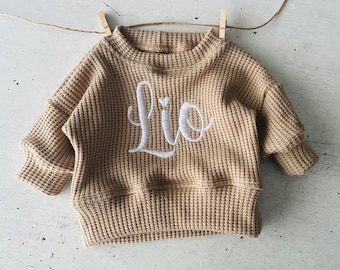May include: A beige waffle knit sweater with long sleeves and a crew neck. The sweater has the name "Lio" embroidered on the front in white.