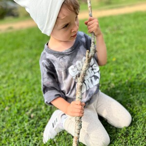 May include: A young child wearing a white beanie with a brown logo and a grey t-shirt with the text "Good Vibes Only" in white. The child is kneeling on green grass and holding a long, brown stick.
