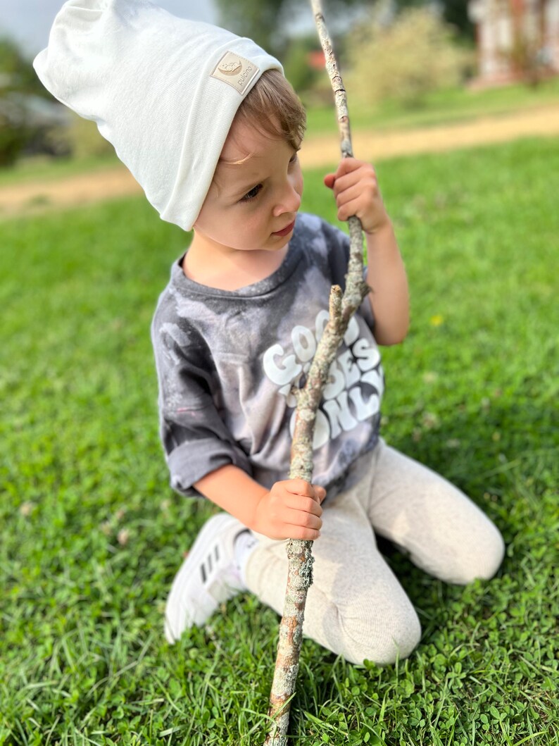 May include: A young child wearing a white beanie with a brown logo and a grey t-shirt with the text "Good Vibes Only" in white. The child is kneeling on green grass and holding a long, brown stick.