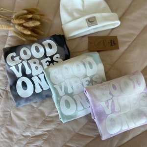 May include: Three tie-dye shirts with the phrase "Good Vibes Only" in white lettering. The shirts are in shades of black, green, and purple.