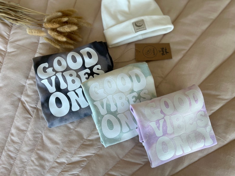 May include: Three tie-dye shirts with the phrase "Good Vibes Only" in white lettering. The shirts are in shades of black, green, and purple.
