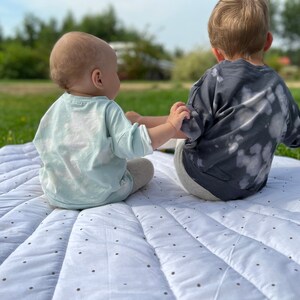 May include: Two children, one wearing a light blue shirt and the other wearing a dark gray shirt with white splotches, sit on a white and black patterned play mat.