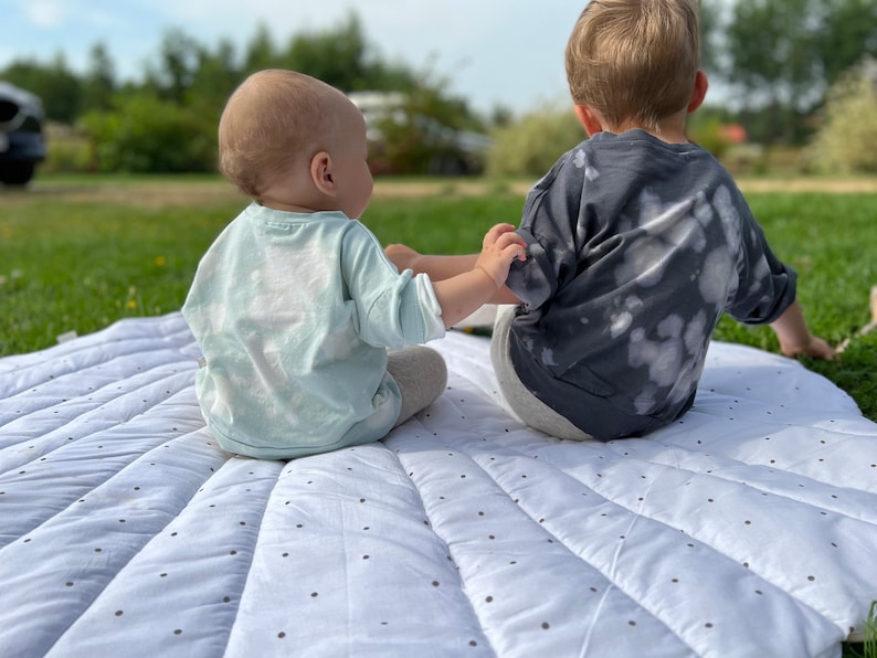 May include: Two children, one wearing a light blue shirt and the other wearing a dark gray shirt with white splotches, sit on a white and black patterned play mat.