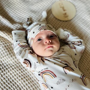Puede incluir: Un bebé con un mono blanco con estampado de arcoíris y estrellas y un gorro anudado marrón y blanco. El mono tiene el texto "Follow your own magic" impreso.