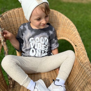 May include: A young child wearing a white beanie with a small logo, a grey t-shirt with the text "GOOD VIBES ONLY", beige leggings, and white sneakers. The child is sitting in a wicker chair.