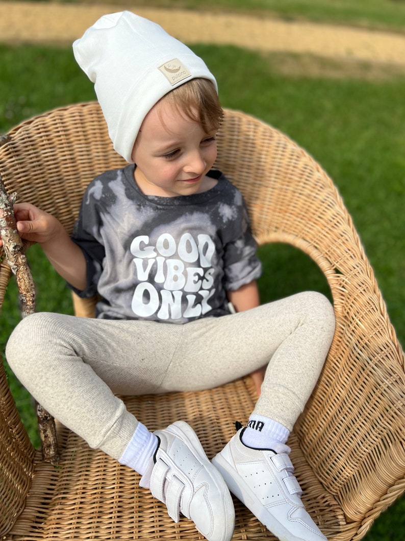 May include: A young child wearing a white beanie with a small logo, a grey t-shirt with the text "GOOD VIBES ONLY", beige leggings, and white sneakers. The child is sitting in a wicker chair.