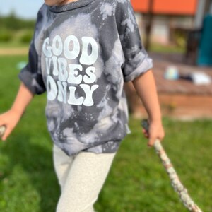 May include: A child wearing a gray tie-dye shirt with the text "GOOD VIBES ONLY" in white. The child is holding a rope and standing on a grassy lawn.