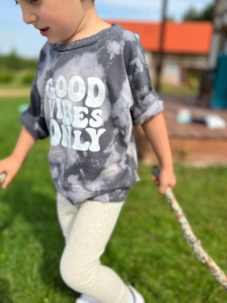 May include: A child wearing a gray tie-dye shirt with the text "GOOD VIBES ONLY" in white. The child is holding a rope and standing on a grassy lawn.