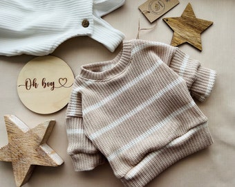 May include: A striped beige and white sweater with long sleeves. The sweater is laid out on a light brown surface with a wooden sign that says "Oh boy" and a wooden star.
