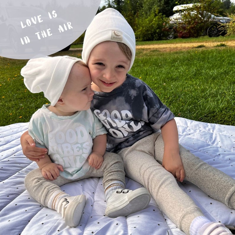 May include: Two children wearing white beanies and matching grey pants sit on a white and grey patterned blanket. The younger child is wearing a light blue shirt with the text "Good Vibes Only" in white. The older child is wearing a dark grey shirt with the text "Good Vibes Only" in white. The text "Love is in the air" is visible in the background.