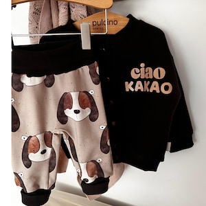May include: A black and brown children's outfit with a brown and white patterned top and black pants. The top has a brown text that says "ciao kakao".