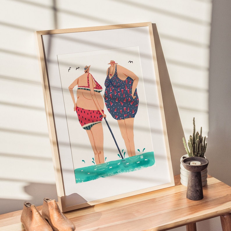 May include: A framed illustration of two women in swimsuits standing in the water. The woman on the left is wearing a red and white watermelon print swimsuit and the woman on the right is wearing a blue and white watermelon print swimsuit. They are both holding a stick.