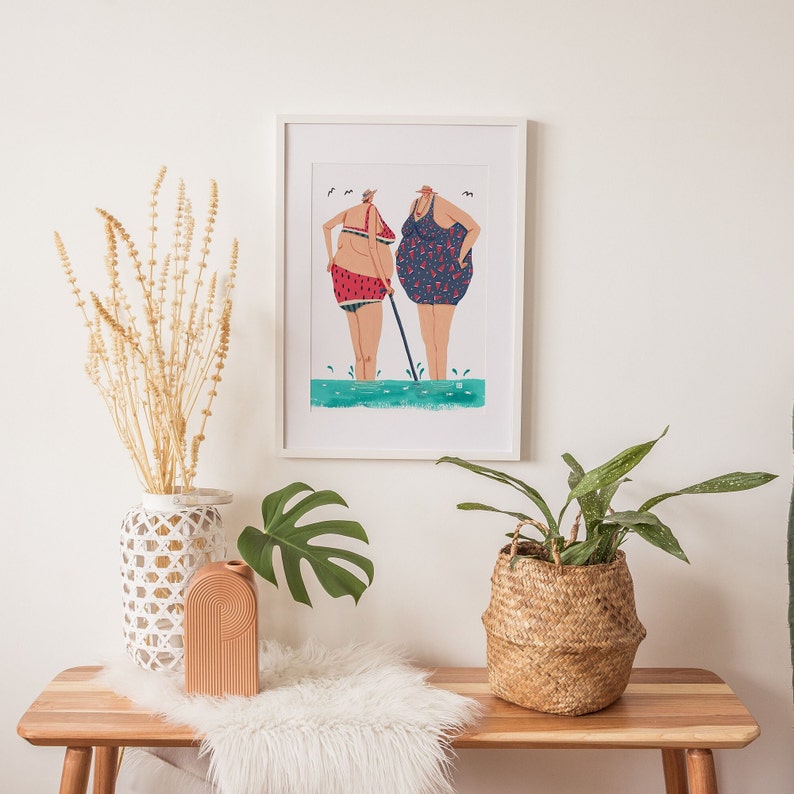 May include: A white framed print of a colorful illustration of two women in swimsuits standing in the water. The women are wearing watermelon print swimsuits and are holding a stick. The print is on a white background and is framed in a white frame.