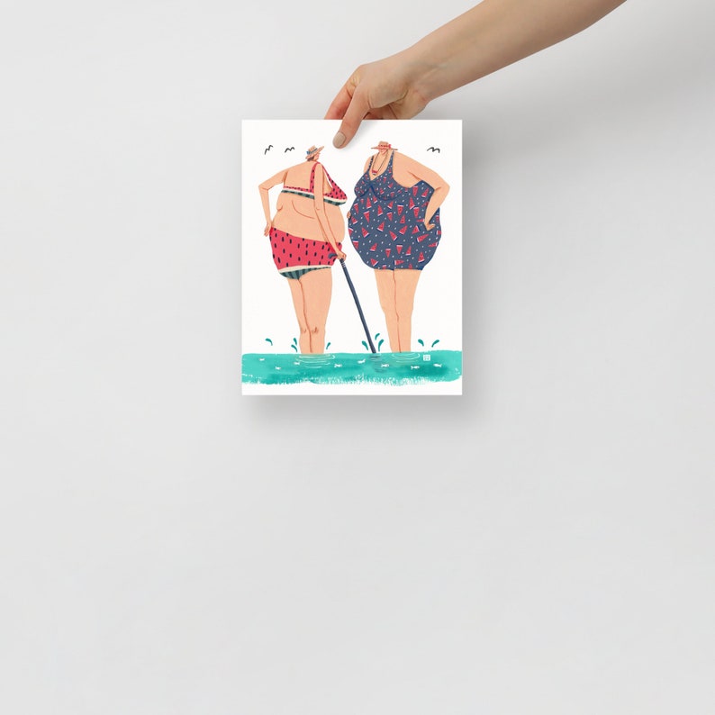 May include: Two women in swimsuits stand in the water. One woman is wearing a red and green watermelon print swimsuit and the other is wearing a blue swimsuit with red and green watermelon print. The woman in the watermelon print swimsuit is holding a stick.