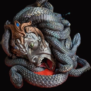 May include: A silver sculpture of a woman's head with snakes wrapped around it. The snakes are black and green with iridescent scales. The woman's mouth is open and her eyes are green.