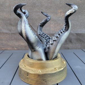 May include: A decorative sculpture featuring octopus tentacles rising from a gold-painted base. The tentacles are silver and grey, with detailed suckers. The base has a metallic gold finish with rivets. A unique decorative item.
