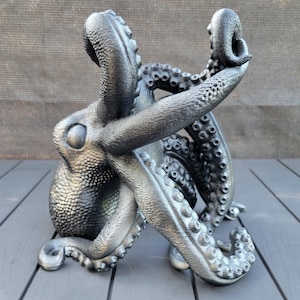 May include: A metallic silver octopus sculpture with detailed textures. The sculpture features multiple tentacles and a detailed head. The octopus is posed as if it is reaching upwards. The sculpture is on a gray surface.
