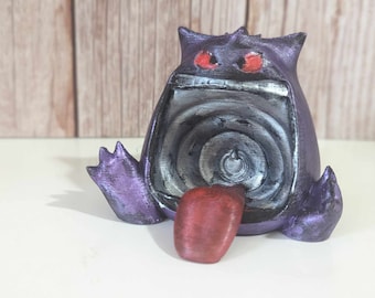 May include: A purple creature figurine with a wide open mouth featuring a metallic silver interior and a protruding red tongue. The figure has two pointed ears, two small arms, and two glowing red eyes set against a dark purple textured body.