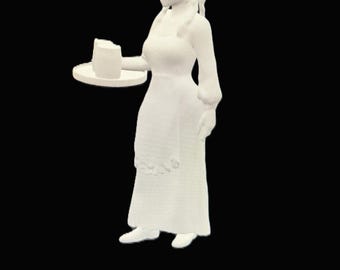 Op de afbeelding: Witte keramische figuur van een vrouw in een lange jurk die een dienblad met een klein taartje vasthoudt.