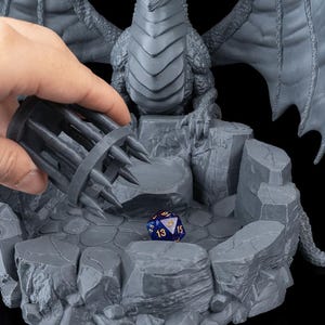 Può includere: Un porta dadi a tema drago grigio con una gabbia e un dado blu e oro. Il drago è appollaiato su una piattaforma rocciosa. La gabbia è tenuta da una mano. Il dado ha i numeri 13, 15 e 5.