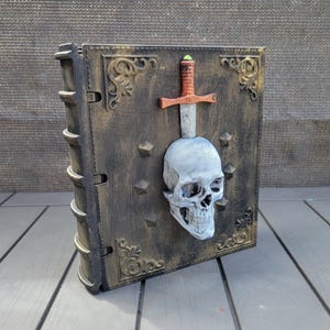May include: A decorative book with a skull and sword design. The book is dark brown with gold accents and a skull pierced by a sword. The spine has a textured design, and the cover features ornate corner details and star-shaped studs.
