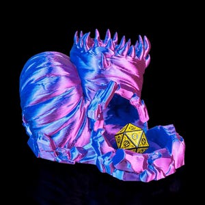 Può includere: Una torre per dadi stampata in 3D, nei toni del blu e del rosa, con una texture rocciosa. Un dado a venti facce giallo e nero è posizionato all'interno. Tema fantasy, per giochi da tavolo.