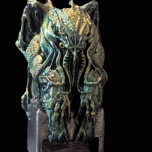 Könnte beinhalten: Eine 3D-gedruckte Figur von Cthulhu, einer fiktiven kosmischen Entität aus den Werken von H.P. Lovecraft. Die Figur ist grün und goldfarben mit einer strukturierten Oberfläche. Sie steht auf einem schwarzen Sockel.