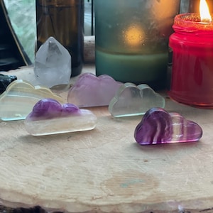 May include: A collection of small, cloud-shaped crystal carvings in various colors, including purple, yellow, and clear. The crystals are arranged on a wooden surface, with candles and other decorative items in the background.