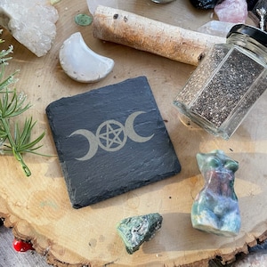 May include: A black slate coaster with a triple moon and pentacle symbol engraved in gold. The coaster is on a wooden surface with other crystals and a glass jar of chia seeds.
