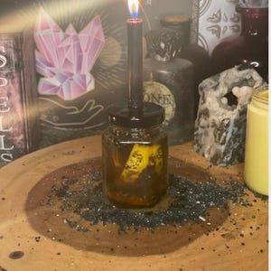 May include: A lit black candle sits atop a small jar filled with a yellow substance and dark particles. The jar is on a wooden surface scattered with black granules. The background includes other jars and a pink crystal illustration.