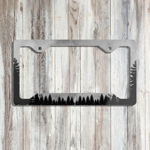 May include: A black metal license plate frame with a silhouette of a forest and a cabin. The frame is designed to fit a standard US license plate.