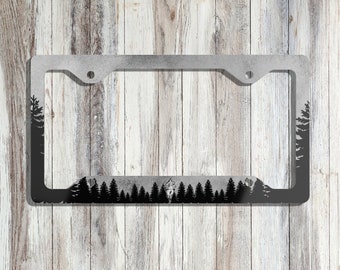 May include: A black metal license plate frame with a silhouette of a forest and a cabin. The frame is designed to fit a standard US license plate.